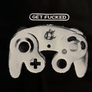 May include: A black fabric surface featuring a white graphic print of a video game controller with a halftone dot pattern. Above the controller, the text GET FUCKED is printed in white inside a rounded rectangular outline.