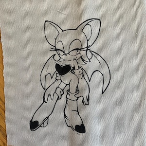 May include: A square piece of light gray fabric with a black line drawing of a cartoon bat-like character. The character has large ears, wings, and is holding a heart. The fabric has a textured appearance.