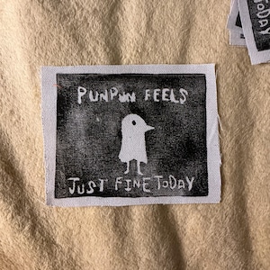 Può includere: Una toppa in tessuto bianco e nero con la scritta "PUNPUN FEELS JUST FINE TODAY" e un'illustrazione di un uccello. La toppa ha un bordo bianco ed è posta su uno sfondo beige.