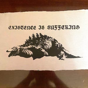 Pode incluir: Gráfico em preto e branco sobre tecido bege claro. O gráfico mostra um monstro estilizado com o texto "EXISTENCE IS SUFFERING" acima. O tecido tem uma borda crua.