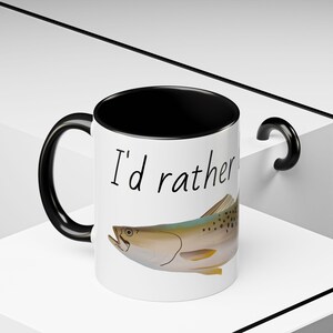 May include: White ceramic mug with a black interior and handle. The mug features the text "I'd rather" and a detailed illustration of a fish. A unique gift for fishing enthusiasts.