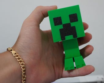 Puede incluir: Una figura verde de Creeper de Minecraft con una cara pixelada negra. La figura se sostiene en una mano, y una pulsera dorada es visible en la muñeca. El Creeper tiene un diseño en bloques.