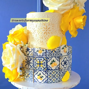 May include: A two-tiered cake decorated with yellow roses and lemon accents. The bottom tier features a blue and white tile design with lemons, while the top tier has a cream-coloured floral pattern. The cake is on a white cake stand. Text: @sweetsformysunshine.