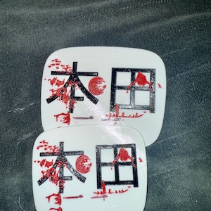 May include: Two white, square coasters with Japanese characters and red accents. The characters are black, with a red sun-like circle and red splatter details. The coasters are stacked on top of each other.
