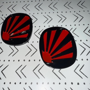Könnte beinhalten: Zwei schwarze, ovale Objekte mit einem roten Sonnenaufgangsdesign. Der Sonnenaufgang zeigt eine rote Sonne und rote Strahlen auf schwarzem Hintergrund. Die Objekte befinden sich auf einer weißen Oberfläche mit einem schwarzen Pfeilmuster.
