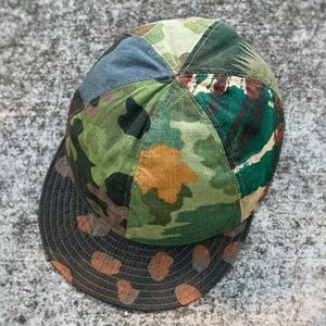 Puede incluir: Gorra de béisbol patchwork con un diseño de camuflaje en tonos verdes, marrones y beige. La visera de la gorra tiene un diseño de lunares marrones y naranjas. La gorra está hecha de varias piezas de tela cosidas.