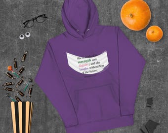 Könnte beinhalten: Ein lila Kapuzenpullover mit einer Vordertasche und Kapuze. Auf der Vorderseite steht der Text "She is clothed in strength and dignity; and she laughs without fear of the future". Zwei orangefarbene Kürbisse und Süßigkeiten sind verstreut.