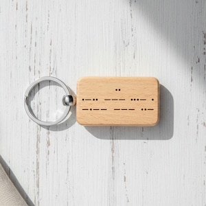 May include: A wooden keychain with a silver key ring. The rectangular wooden tag has Morse code etched into it. The keychain is light brown and sits on a white, textured surface.