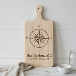 May include: A light-coloured wooden chopping board with a handle, featuring a compass rose design and the text "Bar Harbor, ME" with coordinates. The board is on a white surface.