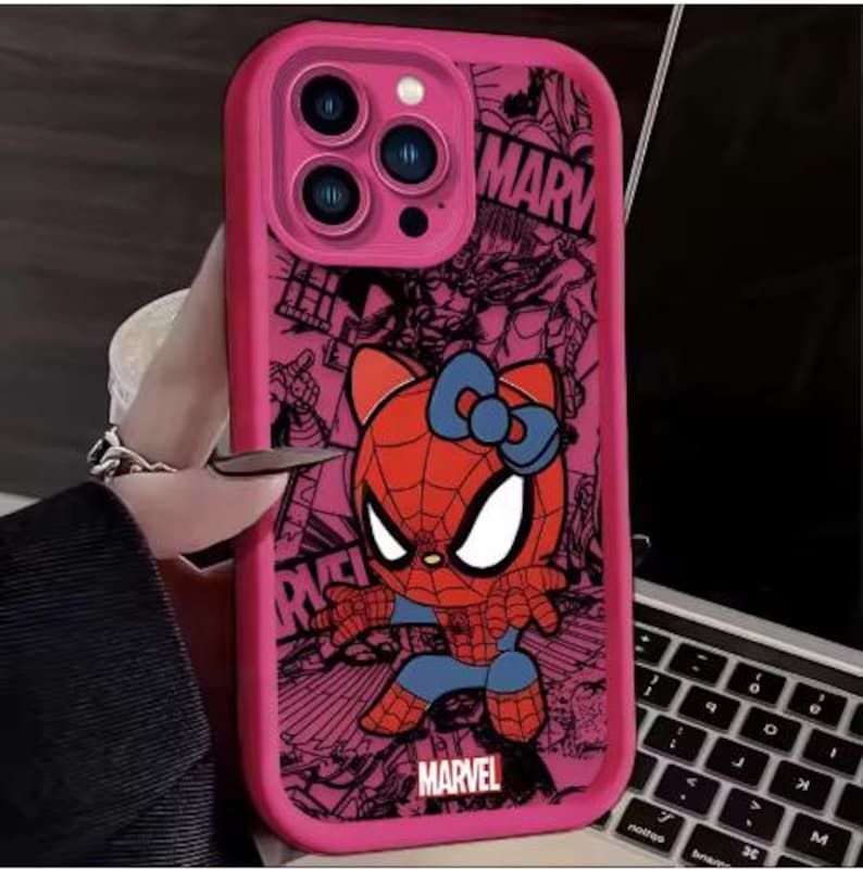 May include: Pink phone case featuring a cartoon Spider-Man character with a cat-like face and a blue bow. The case has a pink border and a Marvel logo. The background has a comic book style design.
