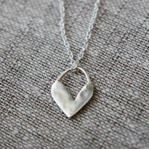 Puede incluir: Un collar de plata con un colgante en forma de corazón. El colgante está abierto en la parte superior y tiene una superficie ligeramente texturizada.