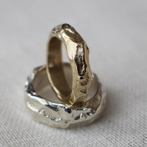 May include: Two textured rings, one gold and one silver, are stacked on a white surface. The gold ring sits atop the silver ring. The rings have an organic, irregular shape, suggesting a handcrafted design. The background is a soft, neutral white.