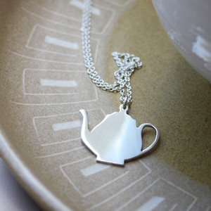 May include: A silver-colored teapot pendant necklace resting on a decorative ceramic plate. The plate is a light brown color with a white geometric pattern. A white teacup is in the background.