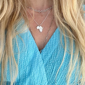 K&ouml;nnte beinhalten: Drei &uuml;bereinander getragene silberne Halsketten. Die oberste Kette ist eine dicke Rundankerkette, die mittlere Kette hat einen kleinen Anh&auml;nger in Form des afrikanischen Kontinents und die untere Kette ist eine lange, d&uuml;nne B&uuml;roklammerkette.