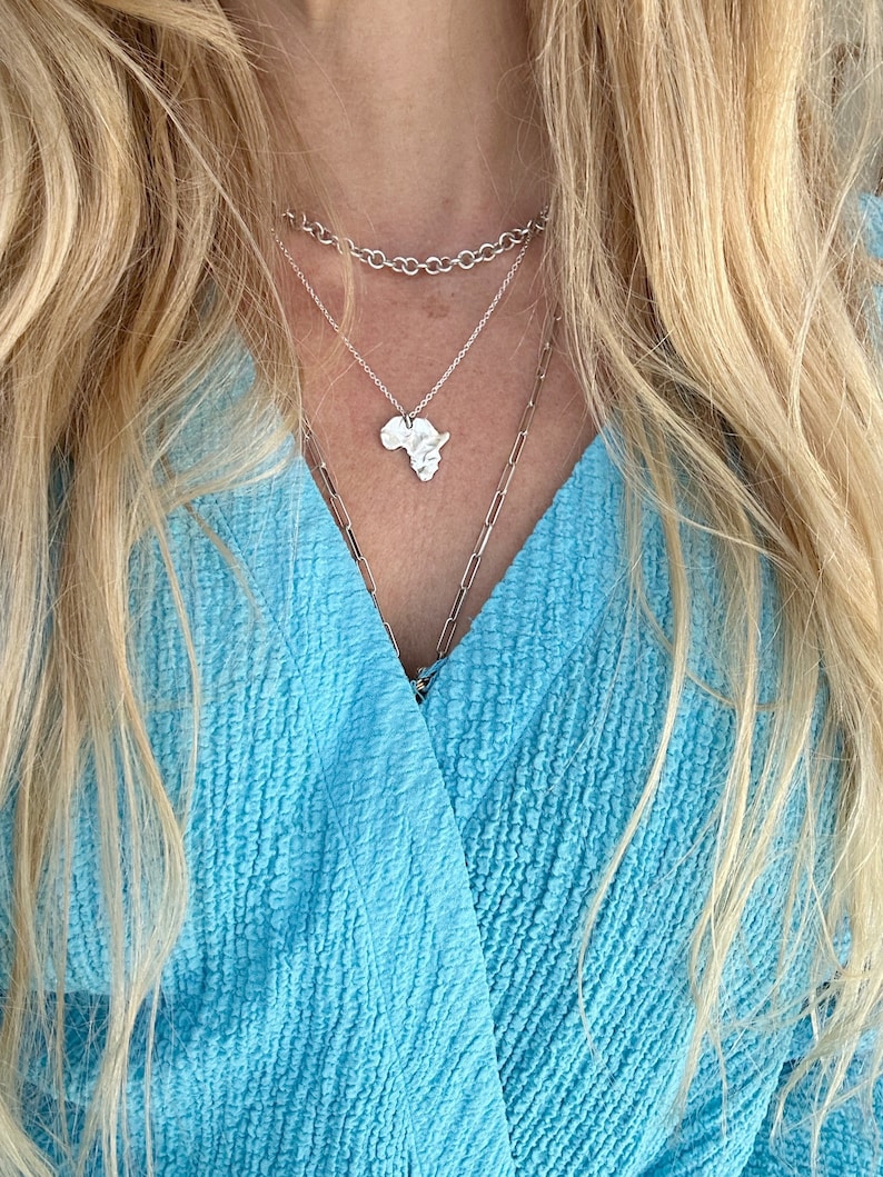K&ouml;nnte beinhalten: Drei &uuml;bereinander getragene silberne Halsketten. Die oberste Kette ist eine dicke Rundankerkette, die mittlere Kette hat einen kleinen Anh&auml;nger in Form des afrikanischen Kontinents und die untere Kette ist eine lange, d&uuml;nne B&uuml;roklammerkette.