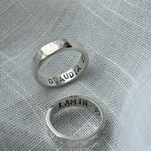May include: Two silver hammered rings with engraved names, "Claudia" and "Lamin", on a white fabric background.