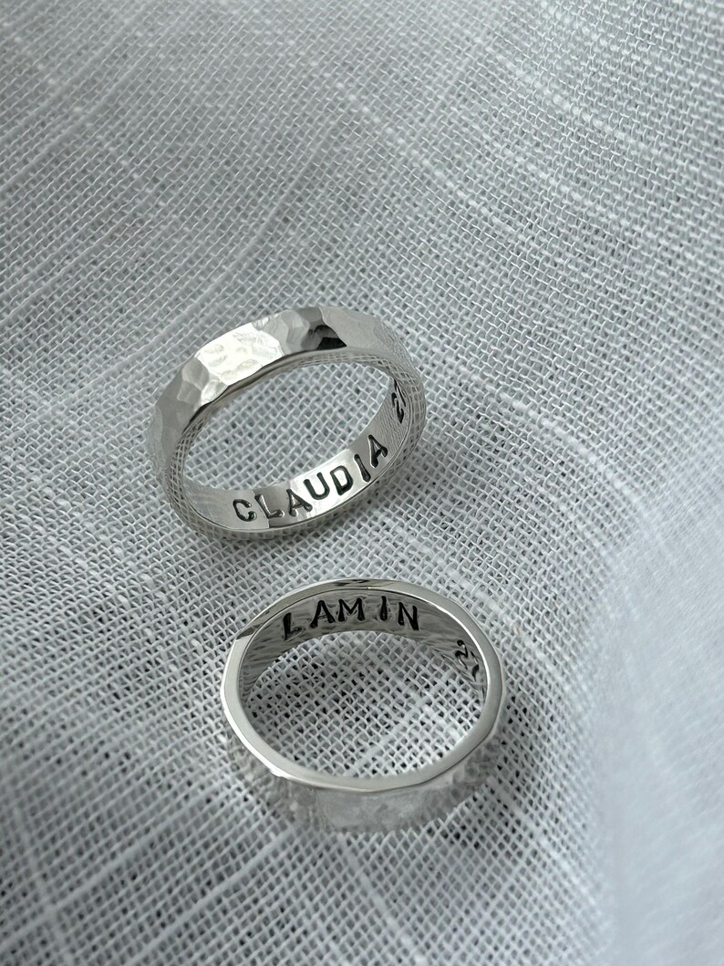 May include: Two silver hammered rings with engraved names, "Claudia" and "Lamin", on a white fabric background.