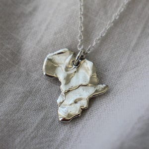 May include: A silver-toned necklace featuring a pendant shaped like the continent of Africa. The pendant has a textured, uneven surface with a metallic sheen and is suspended from a delicate, thin silver-toned chain link necklace.