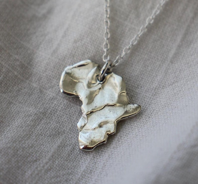 May include: A silver-toned necklace featuring a pendant shaped like the continent of Africa. The pendant has a textured, uneven surface with a metallic sheen and is suspended from a delicate, thin silver-toned chain link necklace.