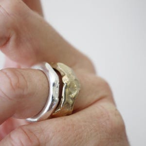 Puede incluir: Dos anillos en un dedo, uno de plata pulida con superficie lisa y redondeada, y otro de oro mate texturizado con forma orgánica irregular y un acabado ligeramente desigual, similar al martillado, en la banda exterior del anillo.