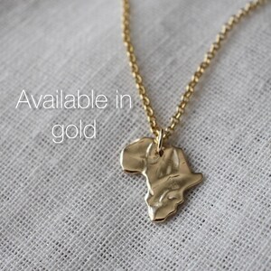 K&ouml;nnte beinhalten: Eine goldfarbene Halskette mit einem Anh&auml;nger in Form des afrikanischen Kontinents mit einer texturierten, geh&auml;mmerten Oberfl&auml;che. Der Anh&auml;nger h&auml;ngt an einer zarten goldfarbenen Kette. Der Text "Available in gold" ist auf dem Bild abgedruckt.