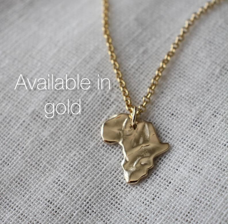 K&ouml;nnte beinhalten: Eine goldfarbene Halskette mit einem Anh&auml;nger in Form des afrikanischen Kontinents mit einer texturierten, geh&auml;mmerten Oberfl&auml;che. Der Anh&auml;nger h&auml;ngt an einer zarten goldfarbenen Kette. Der Text "Available in gold" ist auf dem Bild abgedruckt.