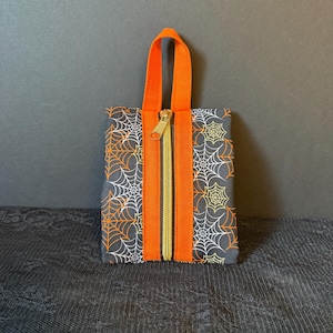 May include: A small, rectangular Halloween-themed bag with an orange handle and zipper. The bag is decorated with a spiderweb pattern in white and orange on a dark gray background. The zipper is gold.