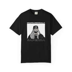 May include: Black t-shirt featuring a graphic of a person with a beard and a patterned head covering. The image is framed by the words "UNAPOLOGETICALLY" above and "SAPERRRA" below. The person in the image is wearing a hat with the initials "SW".