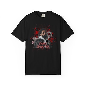 May include: Black t-shirt featuring a graphic of a man with a beard, a snake, and the text "SNAKE CHARMER" in red. The design includes red splatter effects. The shirt is a short-sleeved crew neck.