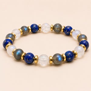 May include: A beaded bracelet featuring alternating round beads of deep blue lapis lazuli, translucent moonstone, and grey labradorite with iridescent flashes. The beads are separated by small, flat, polished gold-tone metal spacers.