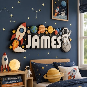 Könnte beinhalten: Ein Kinderzimmer mit Weltraumthema dekoriert. Der Name "JAMES" wird in großen 3D-Buchstaben angezeigt, begleitet von einer Rakete, Planeten und einem Astronauten. Das Zimmer verfügt über sternförmige Wandaufkleber, eine Raketenlampe und Bettwäsche zum Thema Weltraum.