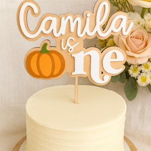 May include: A wooden cake topper with the name "Camila" and the word "one" in white, with a pumpkin graphic. The topper is placed on a frosted cake. The cake is on a wooden cake board.