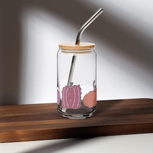 May include: A clear glass can-shaped tumbler with a bamboo lid and a silver metal straw. The glass is decorated with three colorful pumpkins. The tumbler sits on a wooden surface, perfect for fall beverages.