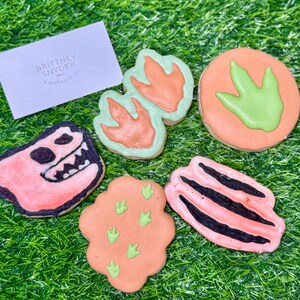May include: A collection of dinosaur-themed cookies with orange, green, and black icing. The cookies are shaped like dinosaur footprints, a dinosaur head, and other dinosaur-related designs. A white card with the text "BRITTNEY SNYDER BAKED ATELIER" is also present.