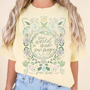 May include: A pale yellow short-sleeve t-shirt featuring a central graphic with the text "the world needs your magic" and "Pixie dust". The design includes green vines, pink flowers, blue accents, a watering can, and sewing supplies like thread.