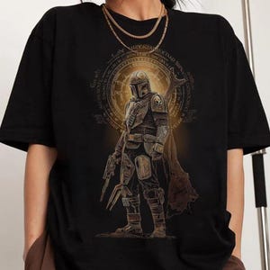 May include: Black t-shirt with a detailed illustration of an armored character holding a weapon, set against a circular design in brown and gold. The shirt is a casual style, suitable for everyday wear.