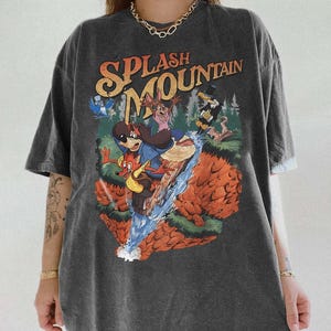 May include: Dark gray t-shirt featuring a colorful graphic of characters on a log flume ride with the text "SPLASH MOUNTAIN" in orange and yellow. The design includes cartoon animals and a waterfall. The shirt has short sleeves and a relaxed fit.