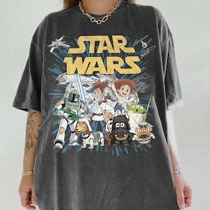 Puede incluir: Camiseta gris oscuro con un dise&ntilde;o de personajes de Star Wars, incluyendo Darth Vader, Woody y Buzz Lightyear. Las palabras "STAR WARS" est&aacute;n en letras amarillas. La camiseta tiene mangas cortas.