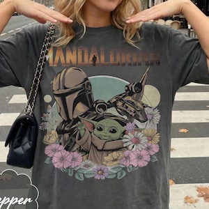 Puede incluir: Camiseta gris oscuro con un dise&ntilde;o de The Mandalorian y Grogu rodeados de flores. El texto "MANDALORIAN" est&aacute; impreso en la parte superior. La camiseta tiene mangas cortas y un corte holgado.