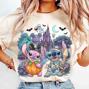 May include: A cream-colored t-shirt featuring a graphic print of two cartoon characters, one dressed as a pumpkin and the other as a mummy, standing in front of a theme park castle, with balloons, bats, and a crescent moon in the background.