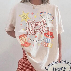 May include: An ivory short-sleeve t-shirt featuring a graphic print with the text "I KNEW IT, I KNEW YOU" and "INFINITY AND BEYOND". The design includes a yellow star, a beaded necklace, a red cowboy boot, a red cowboy hat, and alphabet blocks.