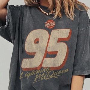 Puede incluir: Camiseta gris oscuro con un gr&aacute;fico desgastado que presenta el n&uacute;mero 95 en crema y rojo, las palabras "Lightning McQueen" y el texto "Rusteze". La camiseta est&aacute; etiquetada como "Comfort Colors Pepper".