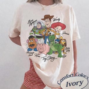 May include: Ivory-coloured t-shirt featuring a graphic of Toy Story characters including Woody, Buzz Lightyear, and Jessie. The shirt has the words "Toy Story" printed below the characters. The shirt is labelled "Comfort Colors Ivory".