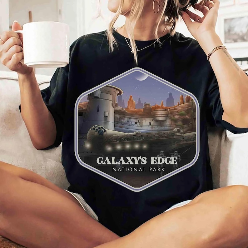 Op de afbeelding: Een zwart t-shirt met een zeshoekige grafische print van een sciencefictionlandschap met een ruimteschip, rotsachtig terrein en twee figuren. De tekst GALAXY'S EDGE NATIONAL PARK staat in witte letters onderaan het ontwerp.