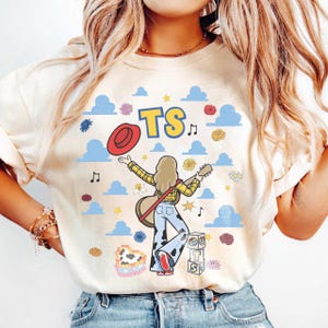 May include: A cream-colored t-shirt featuring a graphic of a person with long hair holding a guitar, wearing a yellow plaid shirt and cow-print pants. The design includes a red hat, blue clouds, flowers, musical notes, and the text TS.