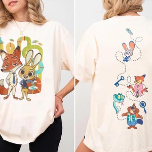 May include: Cream-colored t-shirt featuring cartoon characters of a fox and a rabbit. The front design shows the characters in suits, while the back has a dotted line with other characters. The shirt is a relaxed fit.