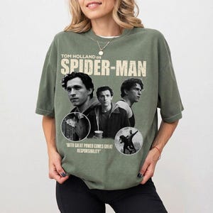 May include: Olive green t-shirt featuring Tom Holland as Spider-Man. The design includes three images of Holland, the words "SPIDER-MAN" in large letters, and the quote "WITH GREAT POWER COMES GREAT RESPONSIBILITY."