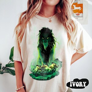 Puede incluir: Camiseta de color marfil con un gr&aacute;fico de un le&oacute;n negro con detalles verdes y tres hienas. El le&oacute;n tiene una expresi&oacute;n amenazante, y el dise&ntilde;o est&aacute; sobre un fondo verde y ahumado. La palabra "IVORY" est&aacute; impresa en negro en negrita.