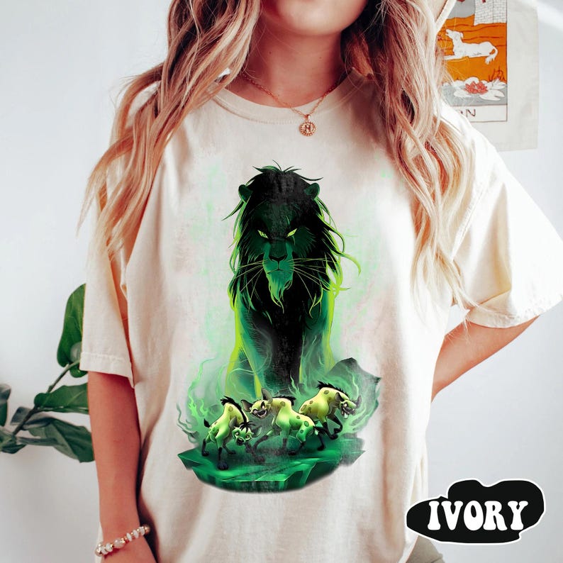 Puede incluir: Camiseta de color marfil con un gr&aacute;fico de un le&oacute;n negro con detalles verdes y tres hienas. El le&oacute;n tiene una expresi&oacute;n amenazante, y el dise&ntilde;o est&aacute; sobre un fondo verde y ahumado. La palabra "IVORY" est&aacute; impresa en negro en negrita.
