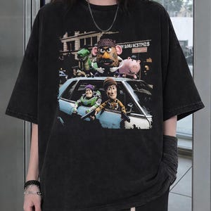 K&ouml;nnte beinhalten: Ein schwarzes T-Shirt mit kurzen &Auml;rmeln und einem Grafikdruck von Toy Story-Figuren. Das Design zeigt eine Gruppe von Charakteren, darunter eine kartoffelf&ouml;rmige Figur, einen gr&uuml;nen Dinosaurier und ein Schwein, die aus einem hellblauen Auto schauen.
